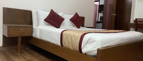 A neatly made bed with a wooden frame, white bedding, and two red pillows. There is a wooden nightstand with a drawer to the left. A wooden door is visible in the background.