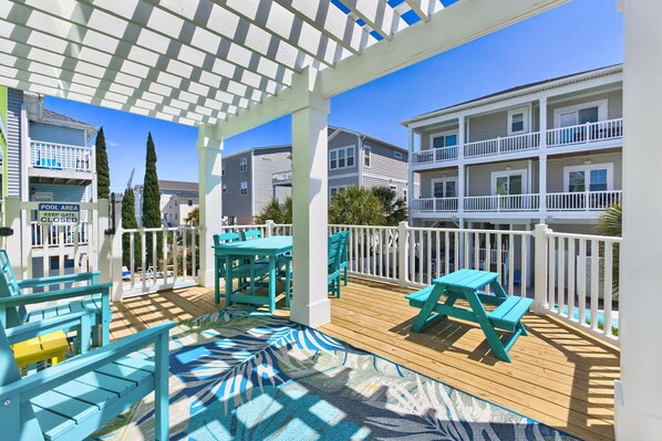 Une terrasse couverte avec des meubles d’extérieur, un panneau indiquant la zone de la piscine, et des immeubles à plusieurs étages en arrière-plan.