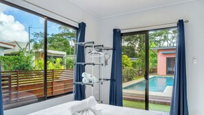 Et hotelværelse med seng, håndklæde, et vindue med udsigt over swimmingpool og grøn bevoksning, samt en balkon med gelænder.