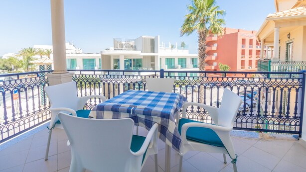 A balcony with a checkered table and white chairs, overlooking a street with buildings and a palm tree.