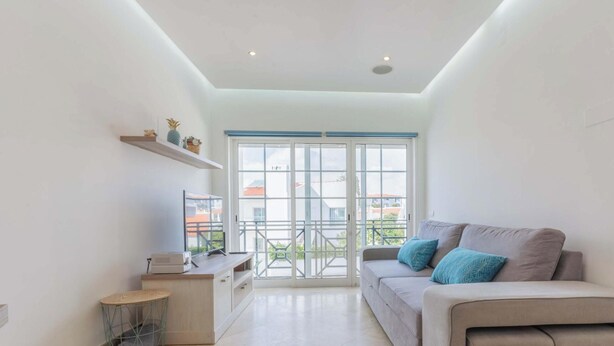 A modern living room with a sofa, a small table, and a TV. There is a large window with a view of buildings outside. - #104 Fanichi Flat by Home Holidays (Albufeira)