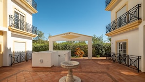 A courtyard with a stone fountain, a covered seating area, and a white building with black railings. - #104 Fanichi Flat by Home Holidays (Albufeira)
