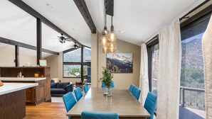 A spacious dining area with a long table and blue chairs, a kitchen with a wooden island, and large windows offering mountain views. - Mtn View, Hot Tub, Open Kitchen, 10m to Sequoia (Three Rivers)