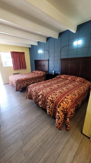 A hotel room with two beds, a wooden floor, a window with red curtains, and a wall with a blue panel.