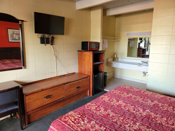 Una habitación de hotel con un aparador de madera, una cama con cubrecama estampado, un televisor montado en la pared, un microondas y un lavamanos con espejo.
