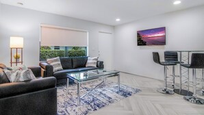 Een moderne woonkamer met een glazen salontafel, donkere banken, een vloerkleed met een blauw patroon, en een stadsgezicht schilderij aan de muur.