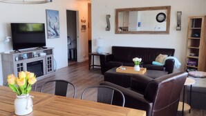 Een woonkamer met een televisie, een houten eettafel met stoelen, een zwarte bank en een houten boekenkast.