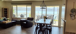 Una sala de estar con comedor, ventanas amplias con vistas a un lago y un balcón con mobiliario exterior.