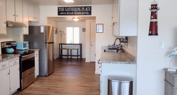 Una cocina moderna con armarios blancos, electrodomésticos de acero inoxidable y encimera de granito. Hay un letrero sobre el fregadero que dice "The Gathering Place where friends become family".