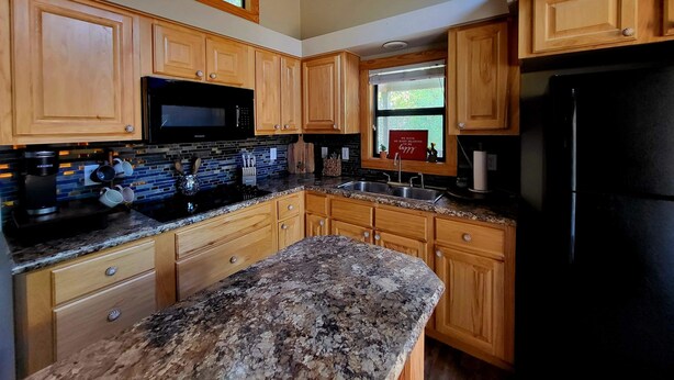 Una cocina con armarios de madera, encimeras de granito, un refrigerador negro y una ventana con vistas a árboles.