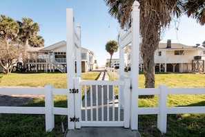 A gated entrance with a white picket fence, leading to a residential area with houses and a palm tree.
