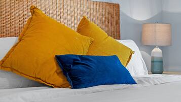 A bed with white sheets and two pillows, one yellow and one blue, in a room with a wicker headboard and a bedside lamp.