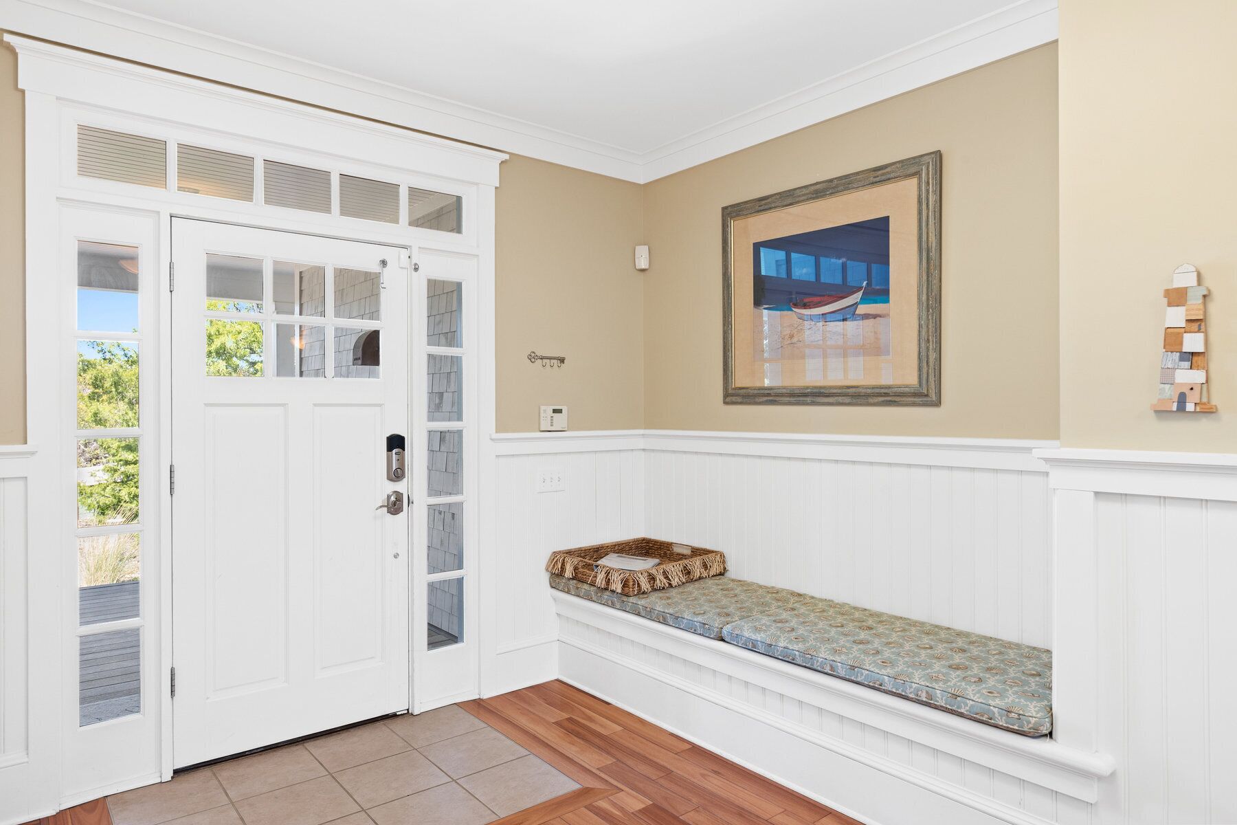 A hallway with a white door, a bench with a cushion, a framed picture on the wall, and a small shelf with decorative items.