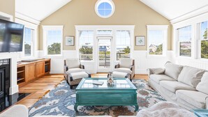 A spacious living room with a high ceiling, a large TV, wooden cabinets, a coffee table, and a view through the glass doors.