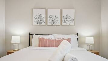 A neatly made bed with white linens, a pink striped pillow, and a rolled white towel. There are two bedside lamps on wooden nightstands. Above the bed, there are three framed black and white floral artworks on the wall.