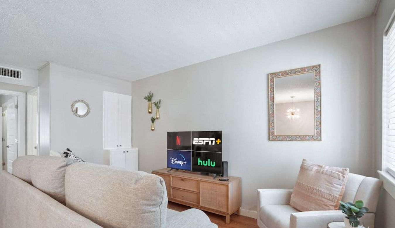Uma sala de estar moderna com um sofá bege, uma poltrona branca e um aparador de madeira com uma TV de tela plana exibindo serviços de streaming.