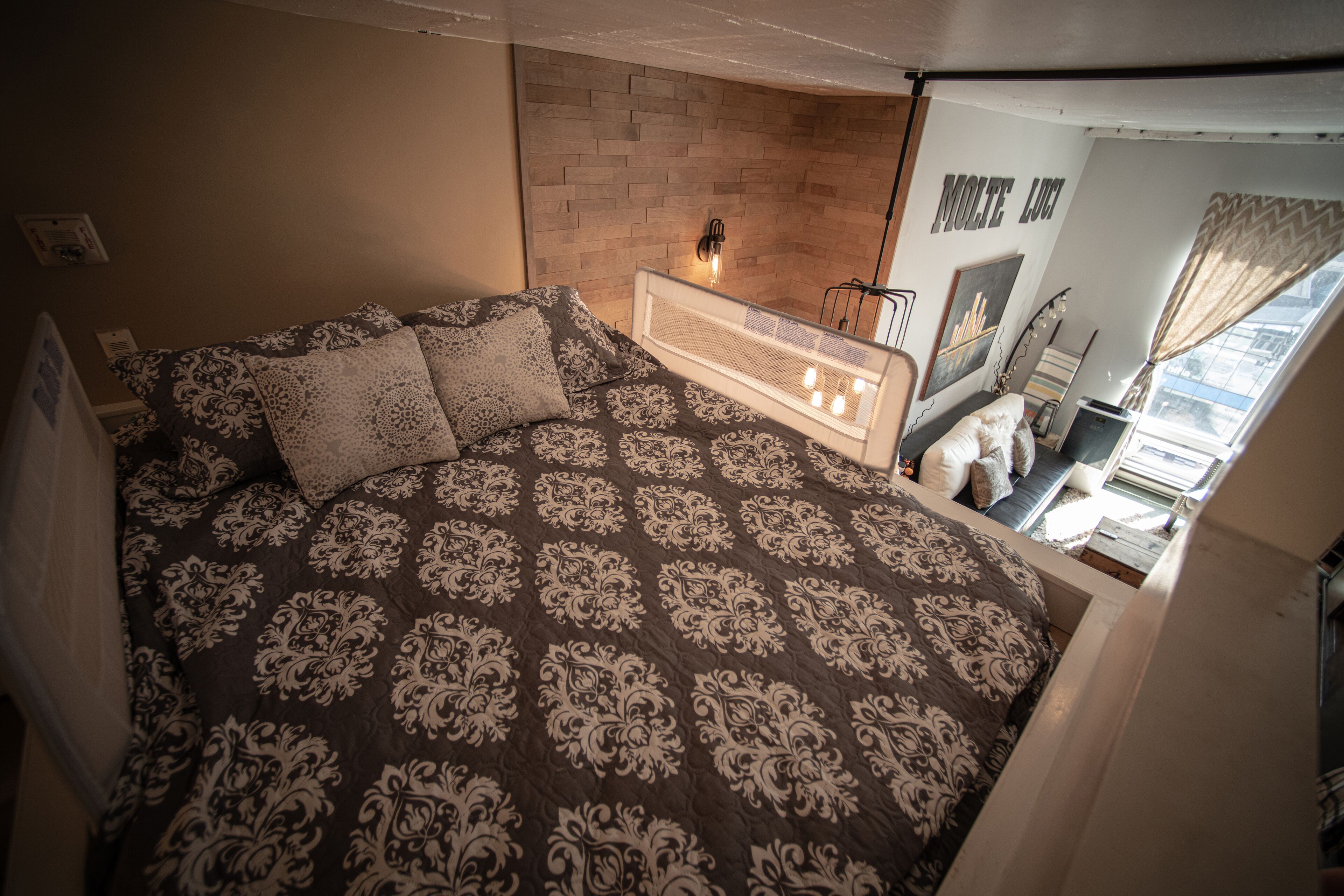 Une chambre à coucher avec une couvre-lit à motifs, un mur en briques, une table de chevet avec une lampe et une fenêtre avec des rideaux.