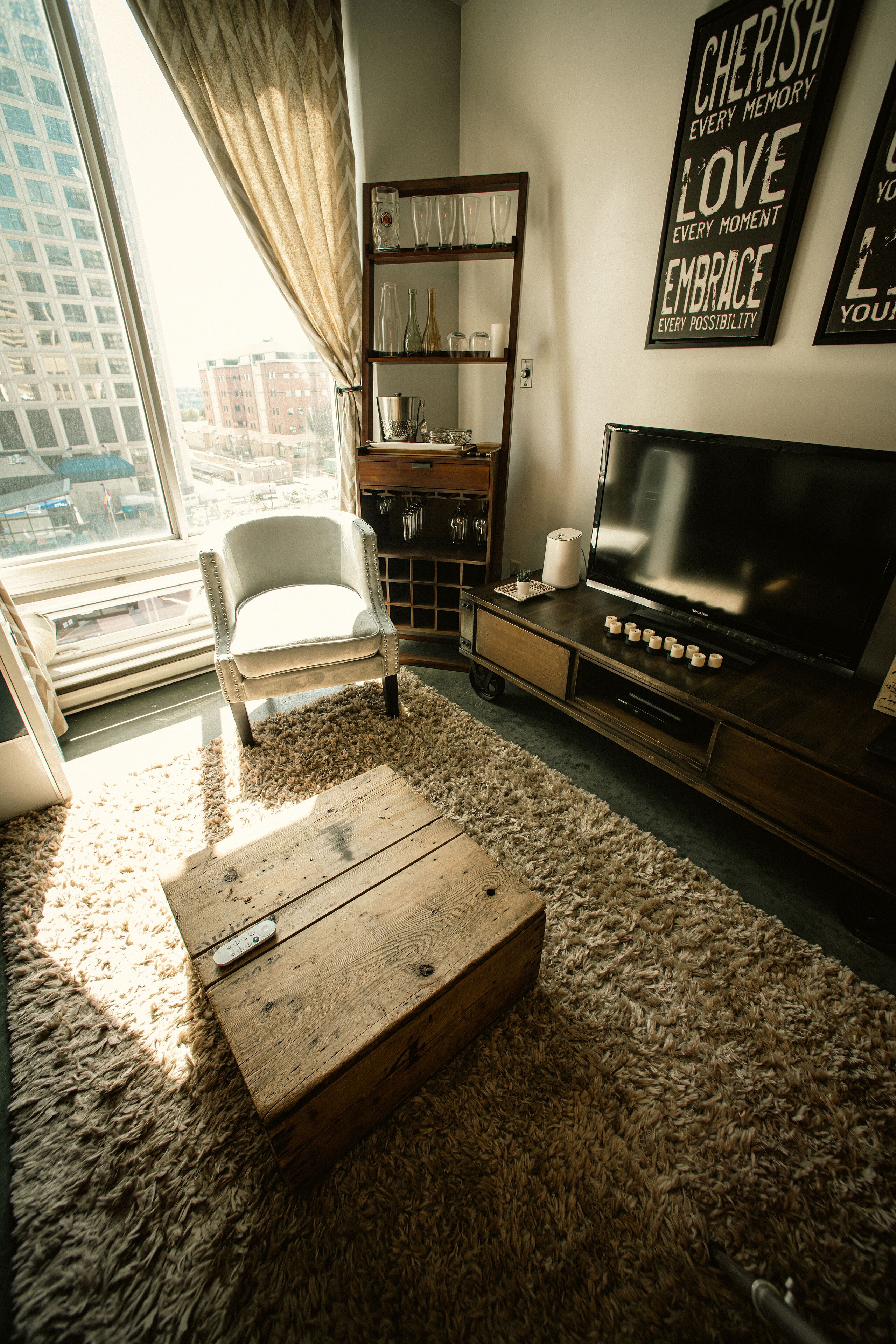 A living room with a large window, a white armchair, a wooden coffee table, a TV, and a shelf with decorative items.