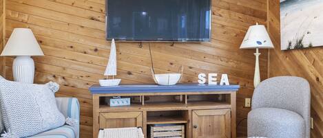 A cozy room with a wooden wall, a flat-screen TV, a sofa, a chair, and a small table with decorative items.