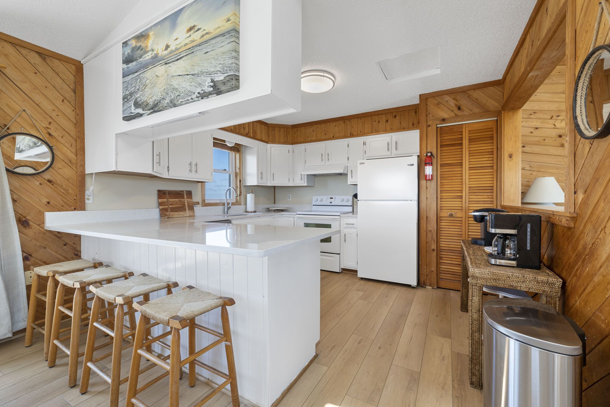 A modern kitchen with white cabinetry, a central island with stools, wooden flooring, and a large painting above the island.