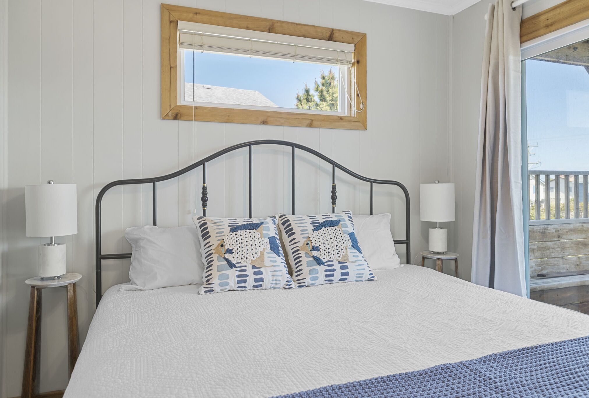 A bedroom with a large bed, two bedside lamps, a window with a wooden frame, and a view of the outdoors.
