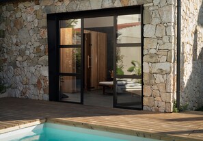 Une maison moderne avec une façade en pierre, une terrasse en bois et une porte vitrée donnant sur un intérieur agrémenté d’un banc en bois et d’une plante.