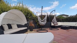 Een terras met een gebogen witte bank, een wit kussenbankje en twee hangmatten.