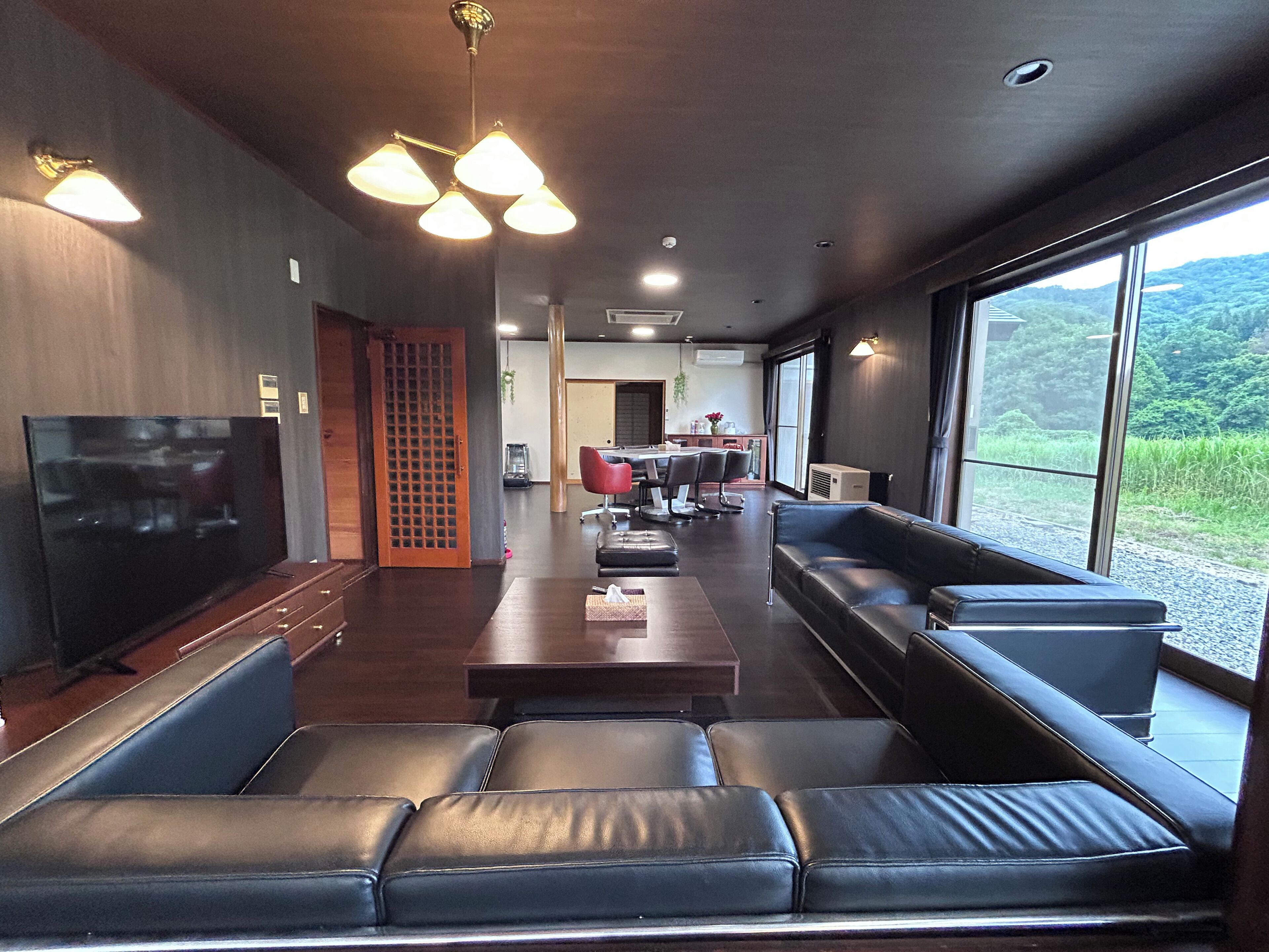 A modern living room with a flat-screen TV, leather sofas, a dining area with chairs, and large windows offering a view of greenery.