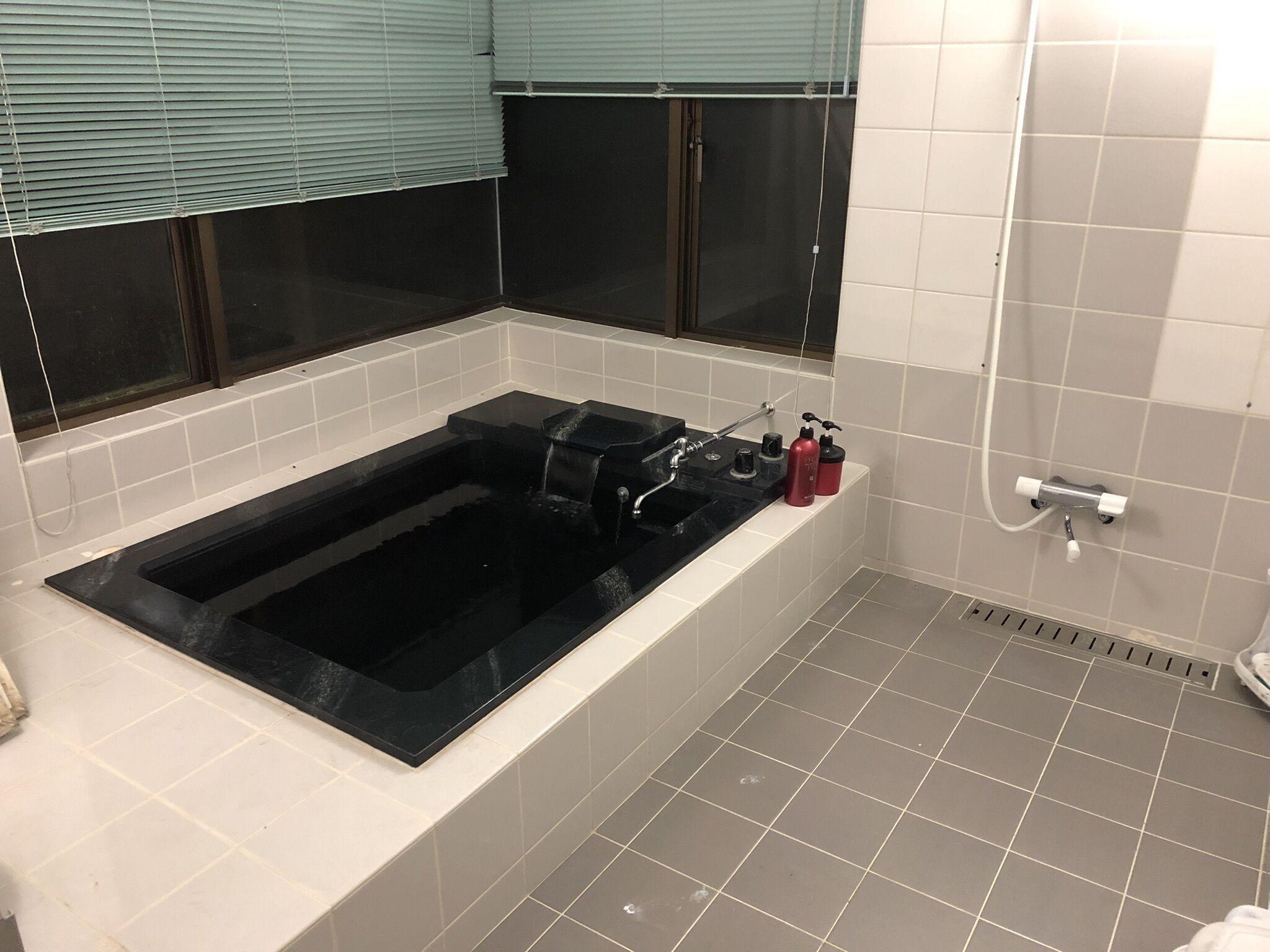 A bathroom with a black bathtub, white tiles, a shower area, and a window with blinds.