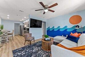 Un salon moderne doté d’un ventilateur de plafond, d’un téléviseur à écran plat, d’un coin repas avec une table et des chaises, et d’une cuisine en arrière-plan.