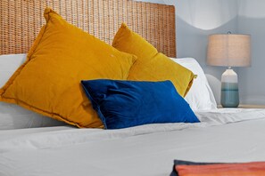 Un lit avec des draps blancs et deux oreillers, un jaune et un bleu, dans une chambre dotée d'une tête de lit en osier et d'une lampe de chevet.