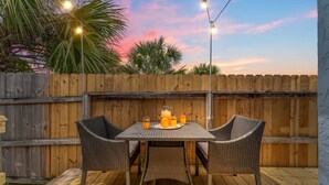 Una terrazza esterna con tavolo in legno e sedie in vimini, una brocca di succo d'arancia e luci a stringa sullo sfondo di una recinzione in legno.