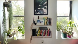 Una habitación con dos ventanas, un cuadro abstracto enmarcado, una estantería con libros y velas, y varias plantas en macetas.