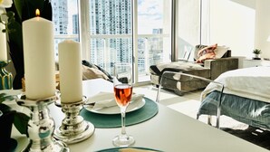 A modern hotel room with a large window offering a city view, a cozy seating area, and a dining setup with candles and a glass of wine. - Perfect Studio 5 Icon Brickell Pool spa gym View (Miami)