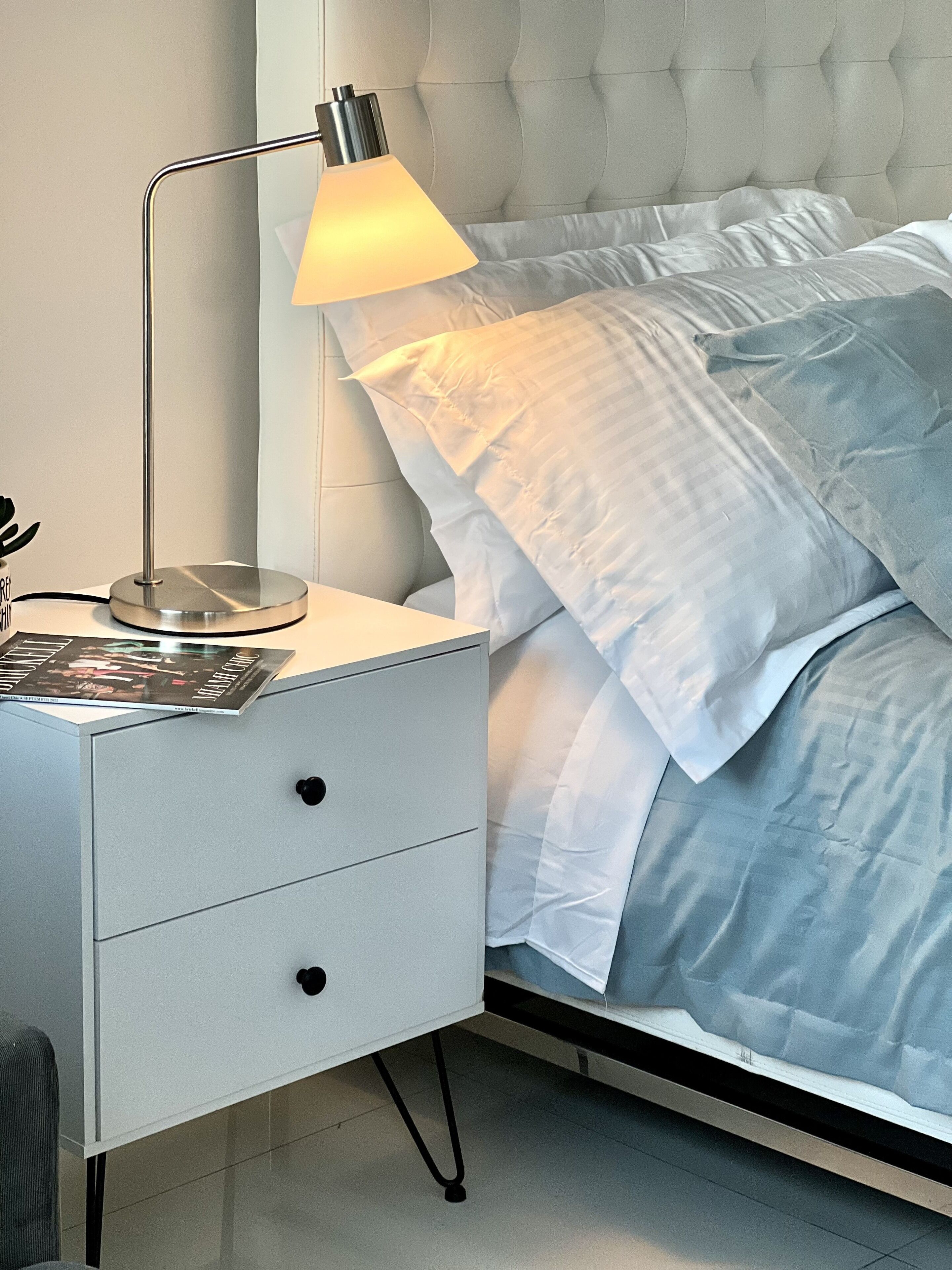 A neatly made bed with a white headboard and pillows, a bedside table with a lamp, and a magazine on the table.
