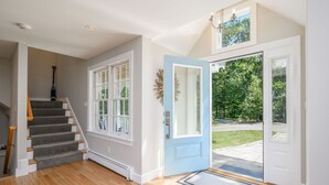 Un hall d’entrée spacieux avec un escalier en bois, une porte bleue et une vue sur un paysage verdoyant.