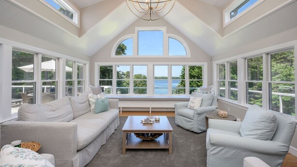 Une pièce spacieuse avec de grandes fenêtres donnant sur un lac, un lustre et un coin salon confortable avec une table basse.