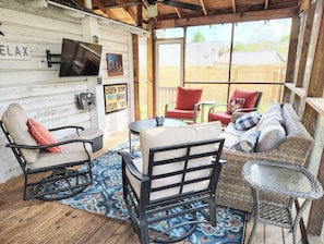 A cozy outdoor seating area with wicker furniture, a TV mounted on the wall, and a wooden deck.