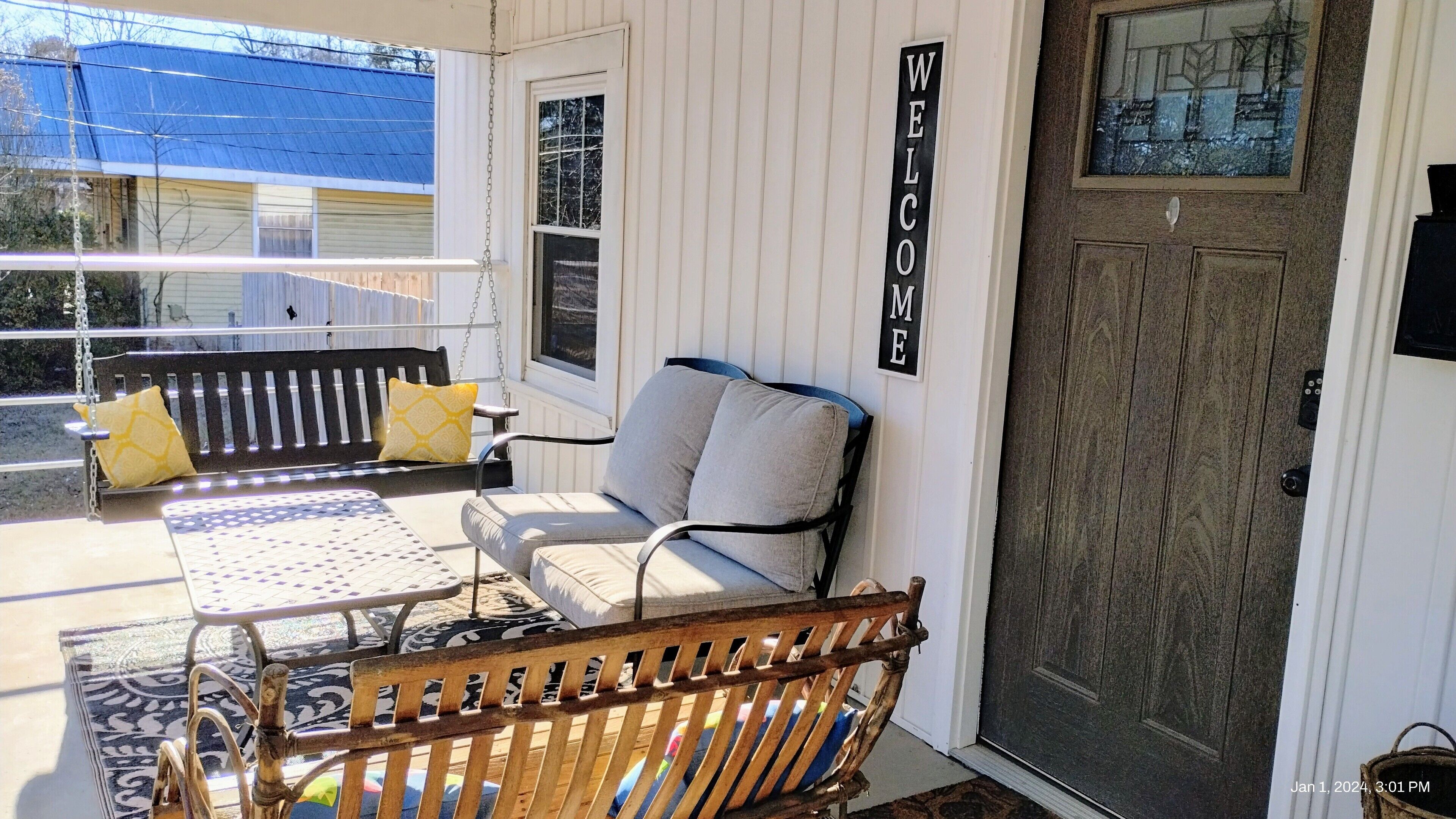 En veranda med bænk, stol og et bord med hynder. Et skilt med ordet 'WELCOME' hænger på væggen.