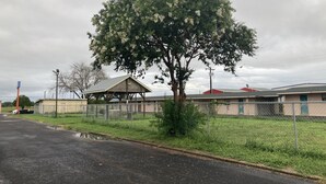 A building with a fenced parking area, a covered structure, and a tree with white flowers. - Capri motel (Alice)