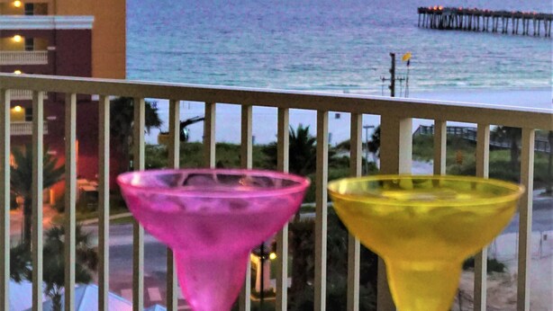 Two colorful cocktail glasses on a round table with a view of the ocean and a pier in the background.