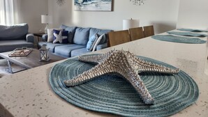 A living room with a starfish decoration on a woven placemat, a sofa with blue cushions, a painting of waves, and a welcome sign.