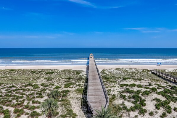 Een strand met een houten looppad dat naar de oceaan leidt, omgeven door duinen en schaarse begroeiing.