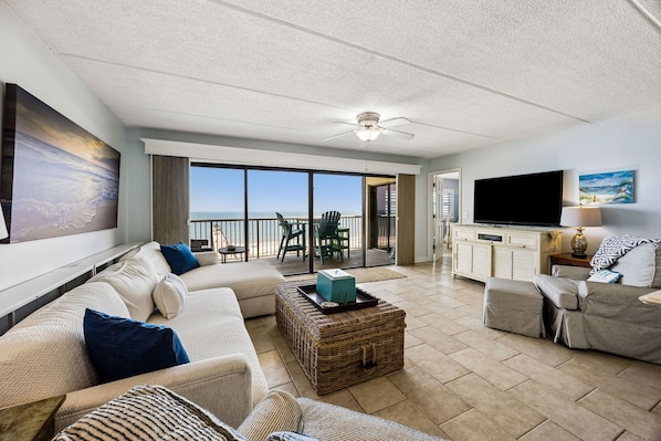 A spacious living room with a large window overlooking a balcony, a flat-screen TV, and a ceiling fan.