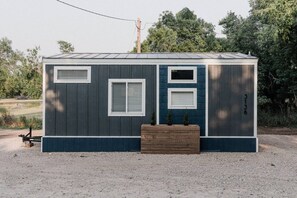 A small, single-story building with a dark exterior, three windows with white frames, and a small wooden planter in front. - Tiny House Creekside · Peaceful Tiny House with great views and wildlife! (Spearfish)