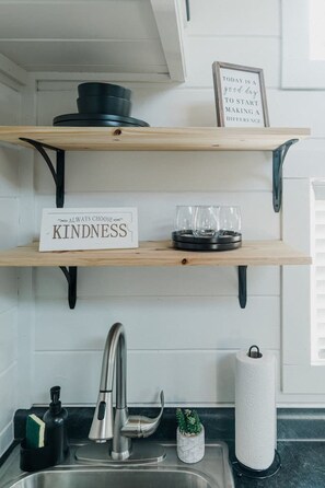 A kitchen with wooden shelves, a sink, and a faucet. There are signs on the shelves, a glass on the middle shelf, and a roll of paper towels on the counter. - Tiny House Creekside · Peaceful Tiny House with great views and wildlife! (Spearfish)