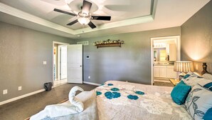 Une chambre à coucher avec un lit, un ventilateur de plafond, une table de chevet, une lampe et une salle de bain visible à travers une porte ouverte.