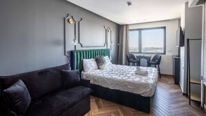 A modern hotel room with a large bed, a dark sofa, a desk, and a window with a city view. - Nissim (Jerusalem)