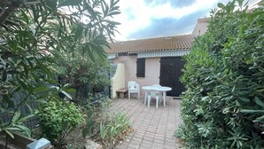 A paved patio with a table and chairs, surrounded by dense foliage and a building with a tiled roof. - Pool pavilion in private residence (Argelès-sur-Mer)