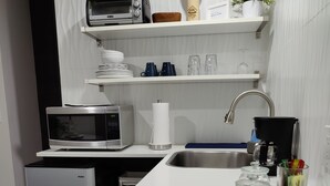 A modern kitchen with a microwave, toaster, and refrigerator. There are shelves above the counter holding dishes and a plant. A sink and faucet are visible, along with a coffee maker and a glass jar with utensils. - 10 Min Brickell & Miami Beach /Parking/Wi-Fi/Grill (Miami)